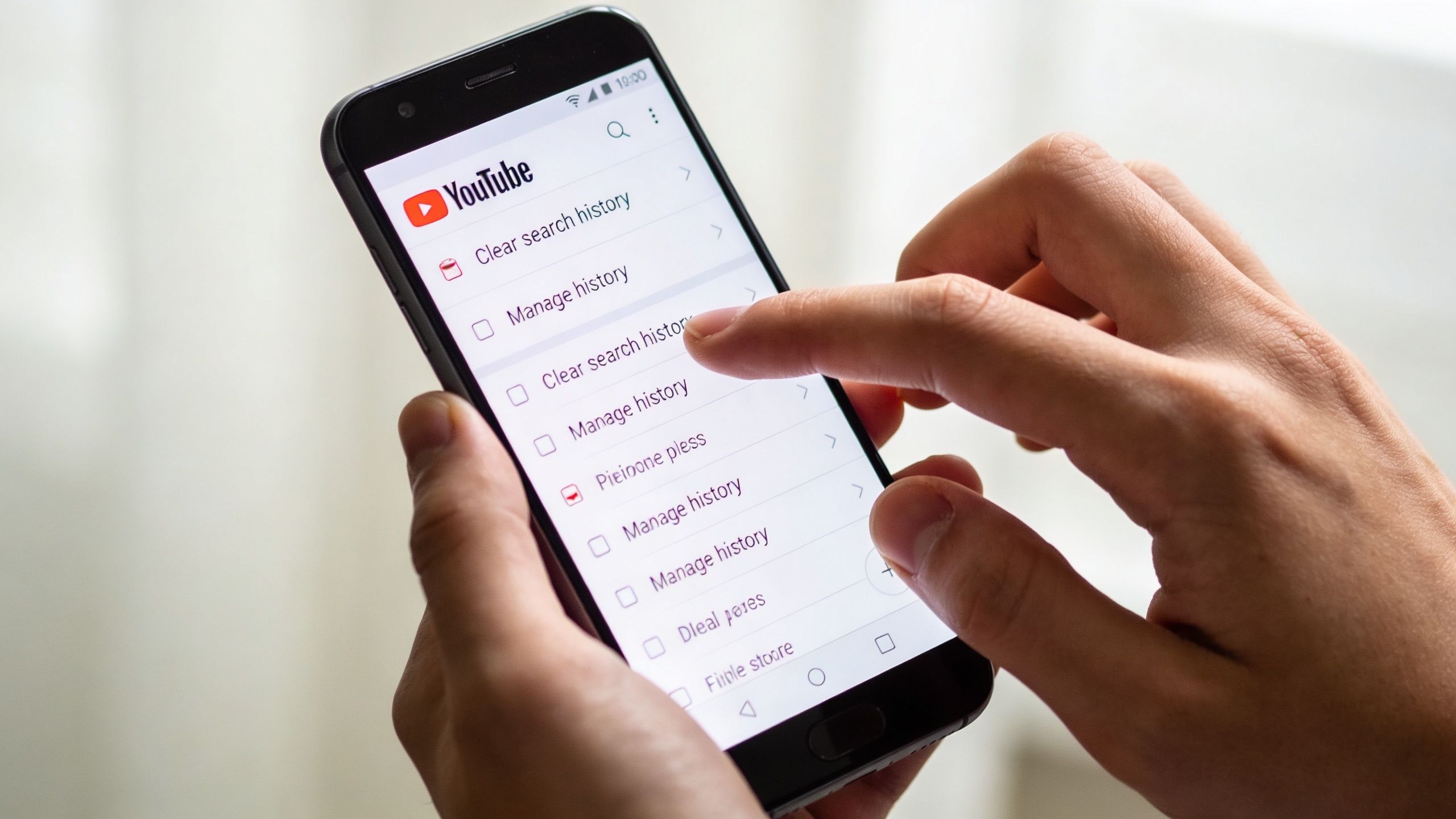 A person using a smartphone to navigate the YouTube application settings menu for clearing search history.